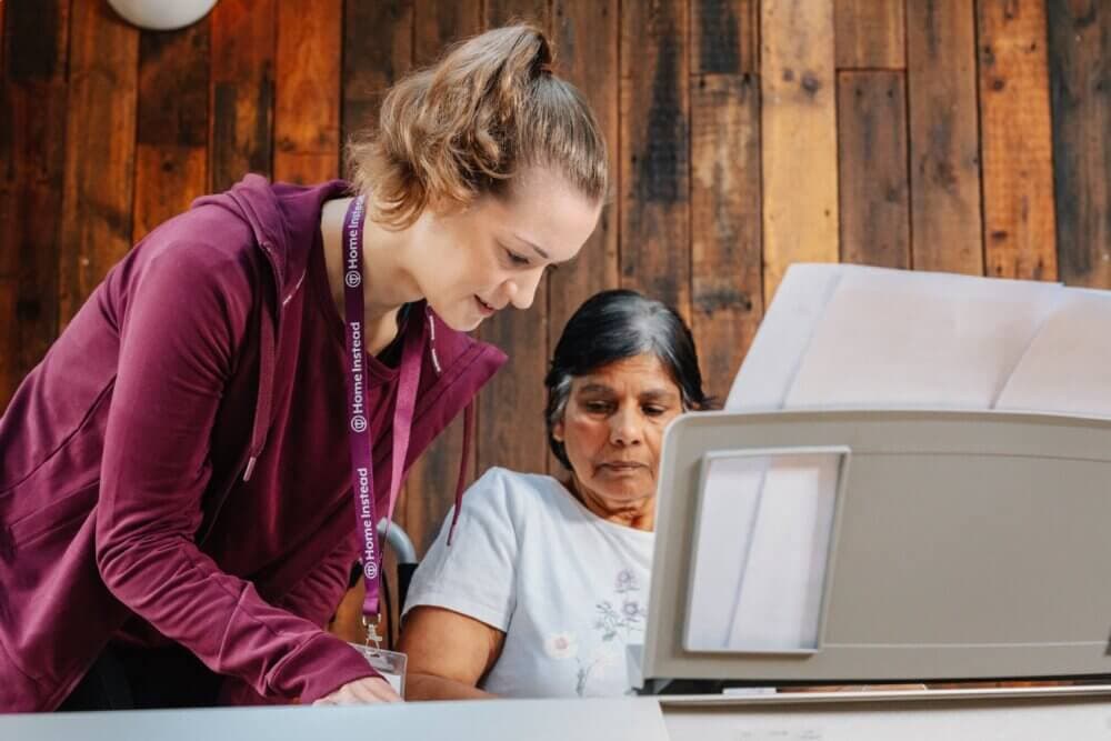 A young woman helps an older woman with paperwork at a desk, in a room with wooden paneled walls. - Home Instead