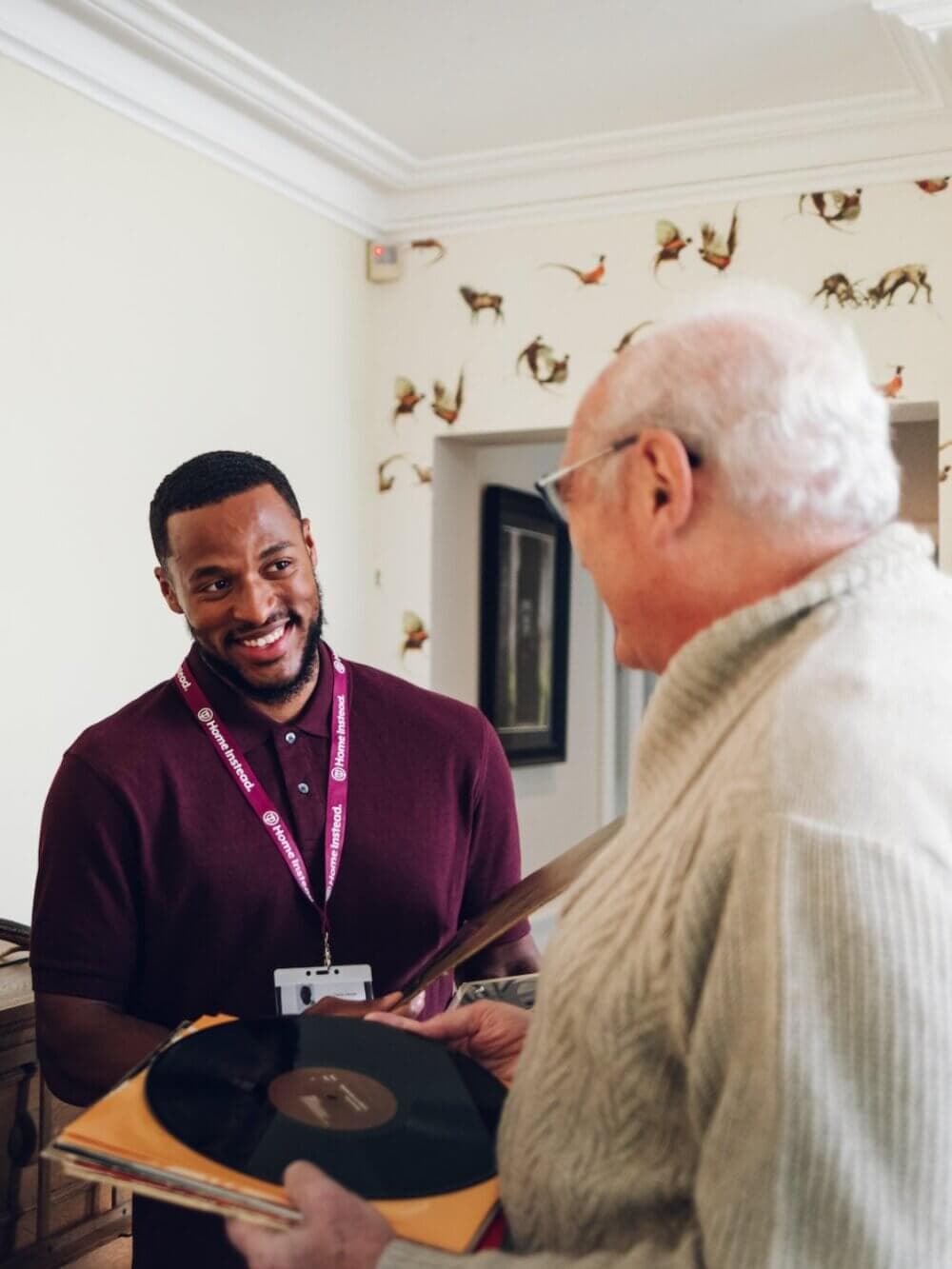A smiling man hands an older man a vinyl record in a warmly lit room with bird-themed wallpaper. - Home Instead