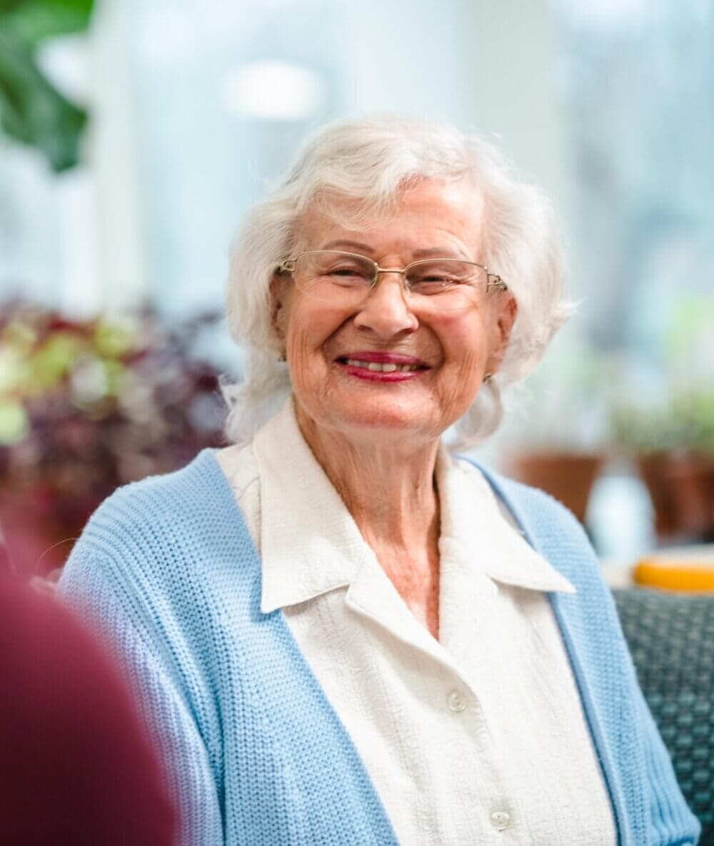Smiling elderly woman with white hair, glasses, and a blue cardigan, sitting indoors with plants in the background. - Home Instead Poole