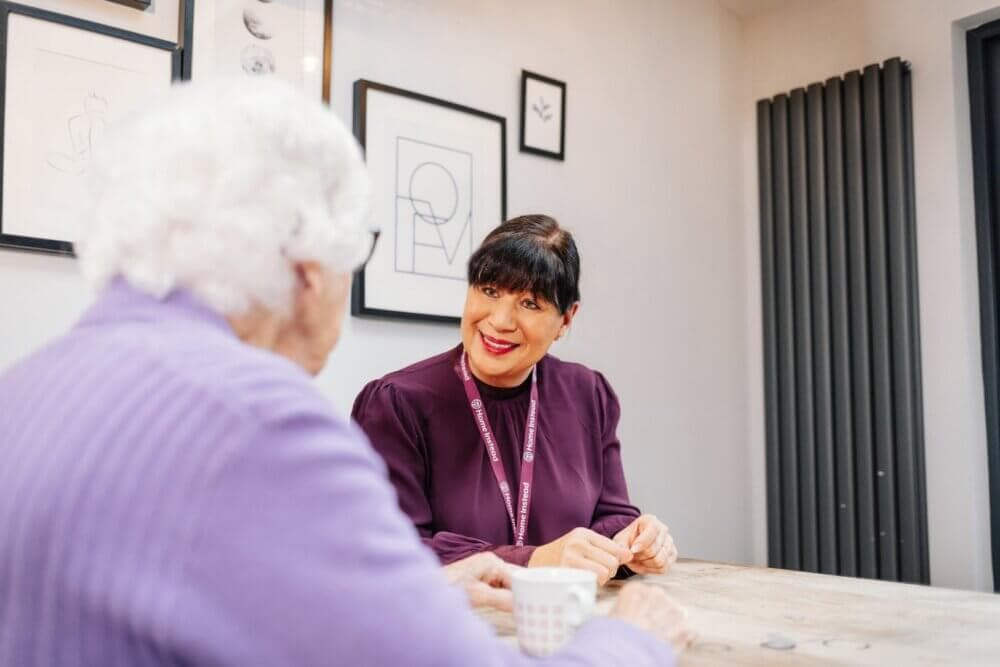 Woman in a purple shirt smiling and talking with an elderly woman over coffee at a table. - Home Instead