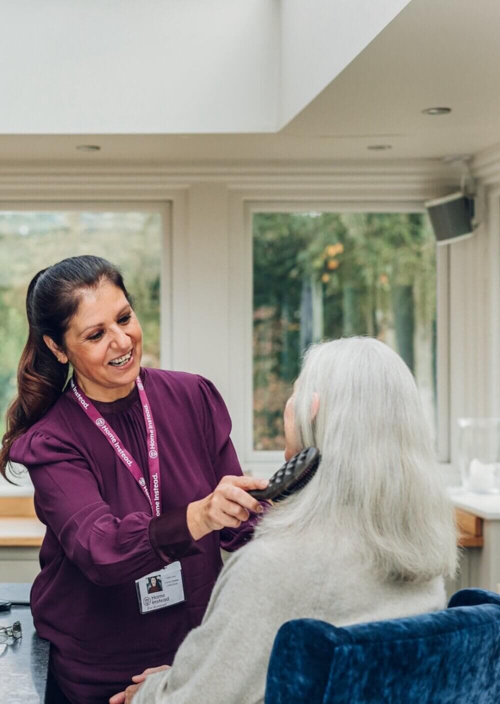 A caregiver in a purple uniform brushes the hair of an elderly woman with gray hair in a bright room. - Home Instead
