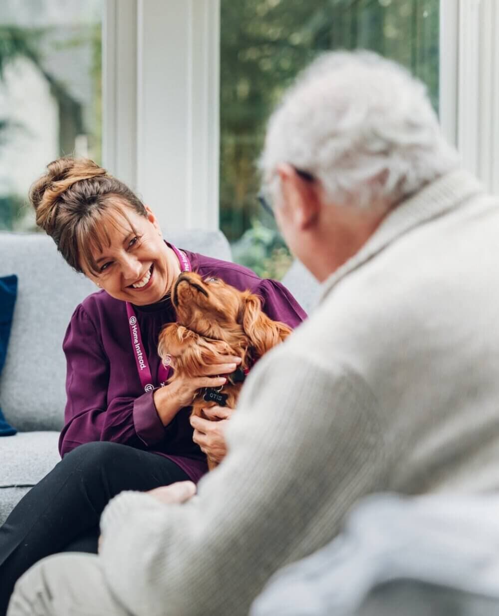 A woman smiles, holding a dog, while sitting across from an elderly man in a cozy, light-filled room. - Home Instead