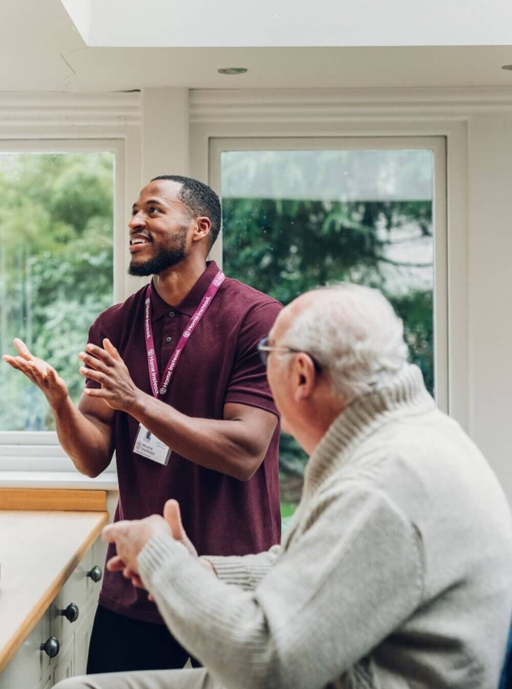A caregiver in a maroon shirt talking and clapping his hands, while an elderly man listens in a cozy room with large windows. - Home Instead