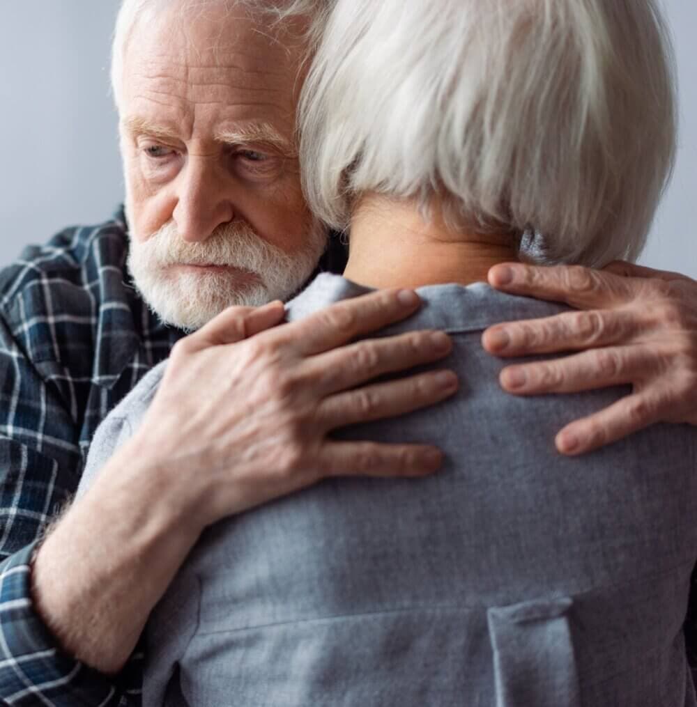 Elderly man with white hair and beard hugging an elderly woman with short white hair, conveying a sense of comfort and support. - Home Instead