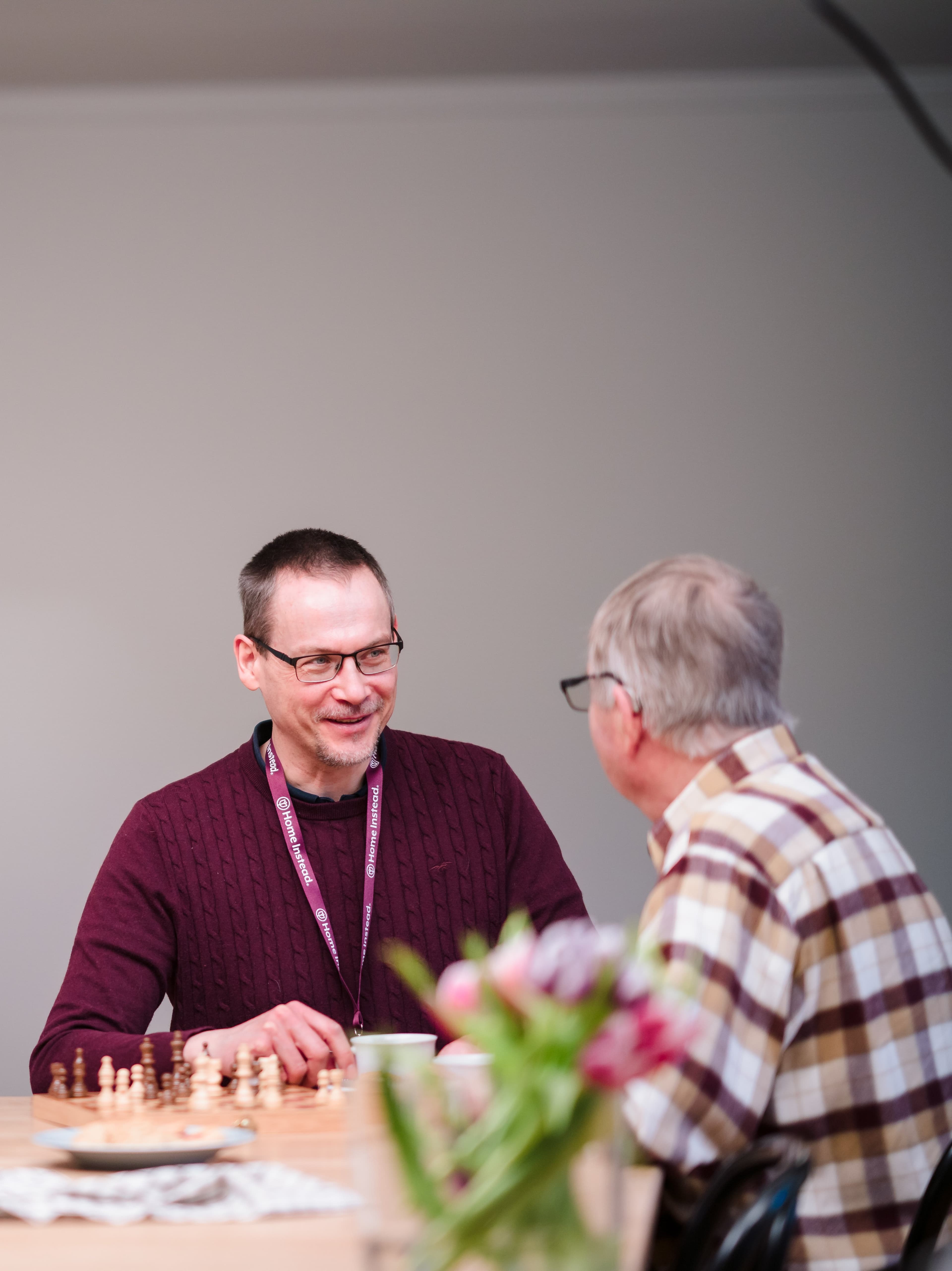 Two men chatting at a table with a chessboard and flowers in the foreground. - Home Instead