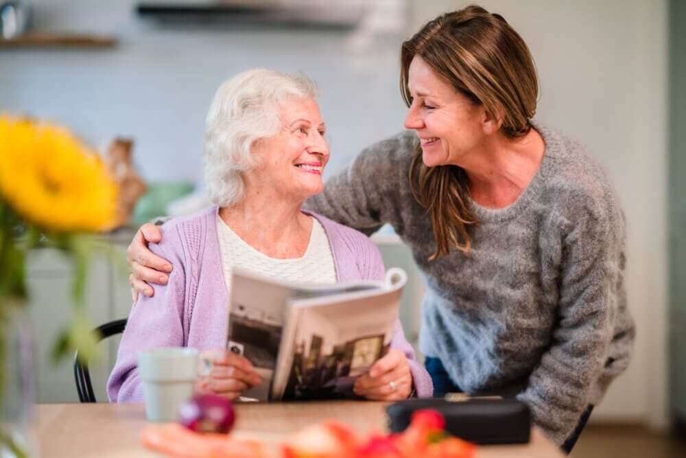 An elderly woman reading a book and smiling, while a younger woman stands beside her, embracing her shoulders. - Home Instead Southampton