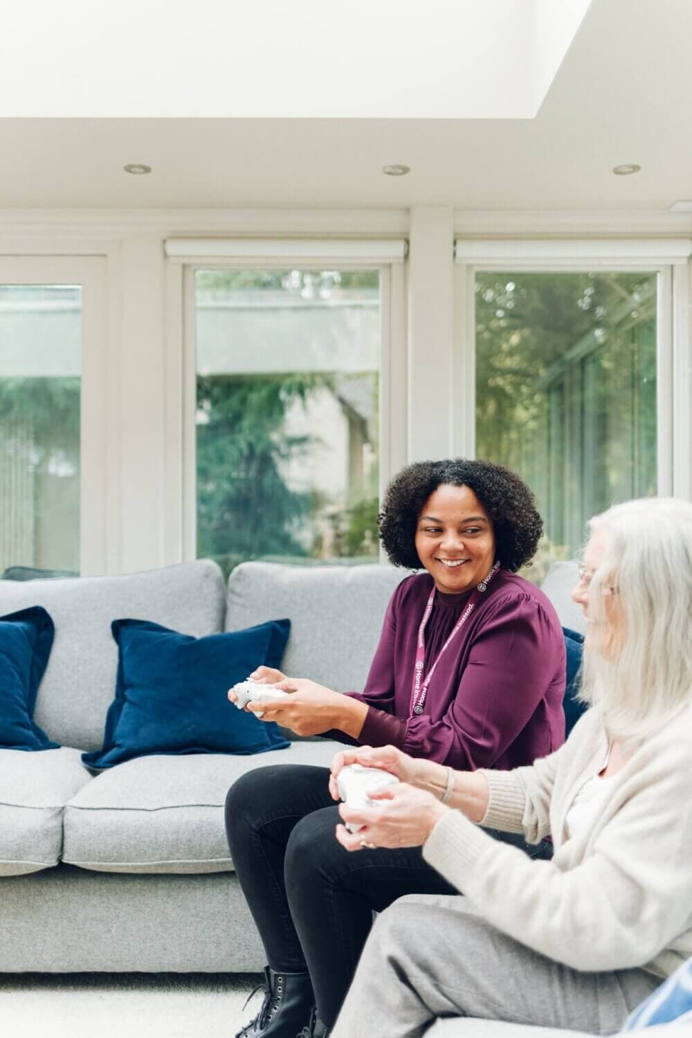 Two women sitting on a sofa, smiling and playing video games together in a bright, modern living room. - Home Instead