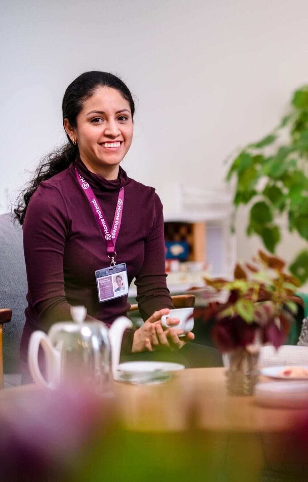 Smiling woman with a name tag sitting in a cozy room, holding a cup of tea, with a teapot and plant in the foreground. - Home Instead