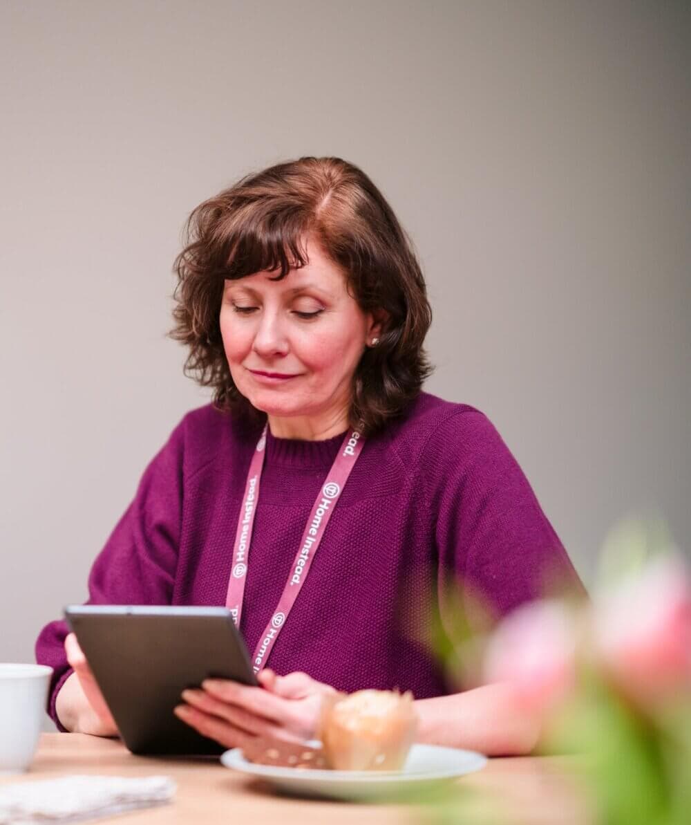 A woman wearing a purple sweater and a lanyard reads a tablet at a table with a cup and a plate with a bun. - Home Instead
