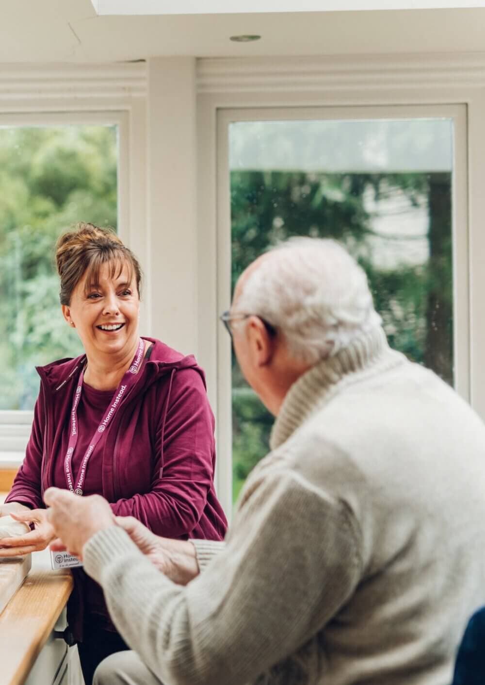A caregiver in a maroon top smiles and talks with an elderly man wearing glasses and a gray sweater at a table. - Home Instead