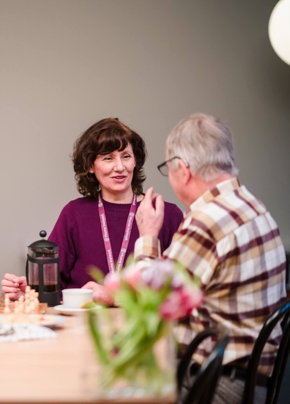 A woman with a French press converses with an older man, both seated at a table with a blurred vase of flowers in the foreground. - Home Instead