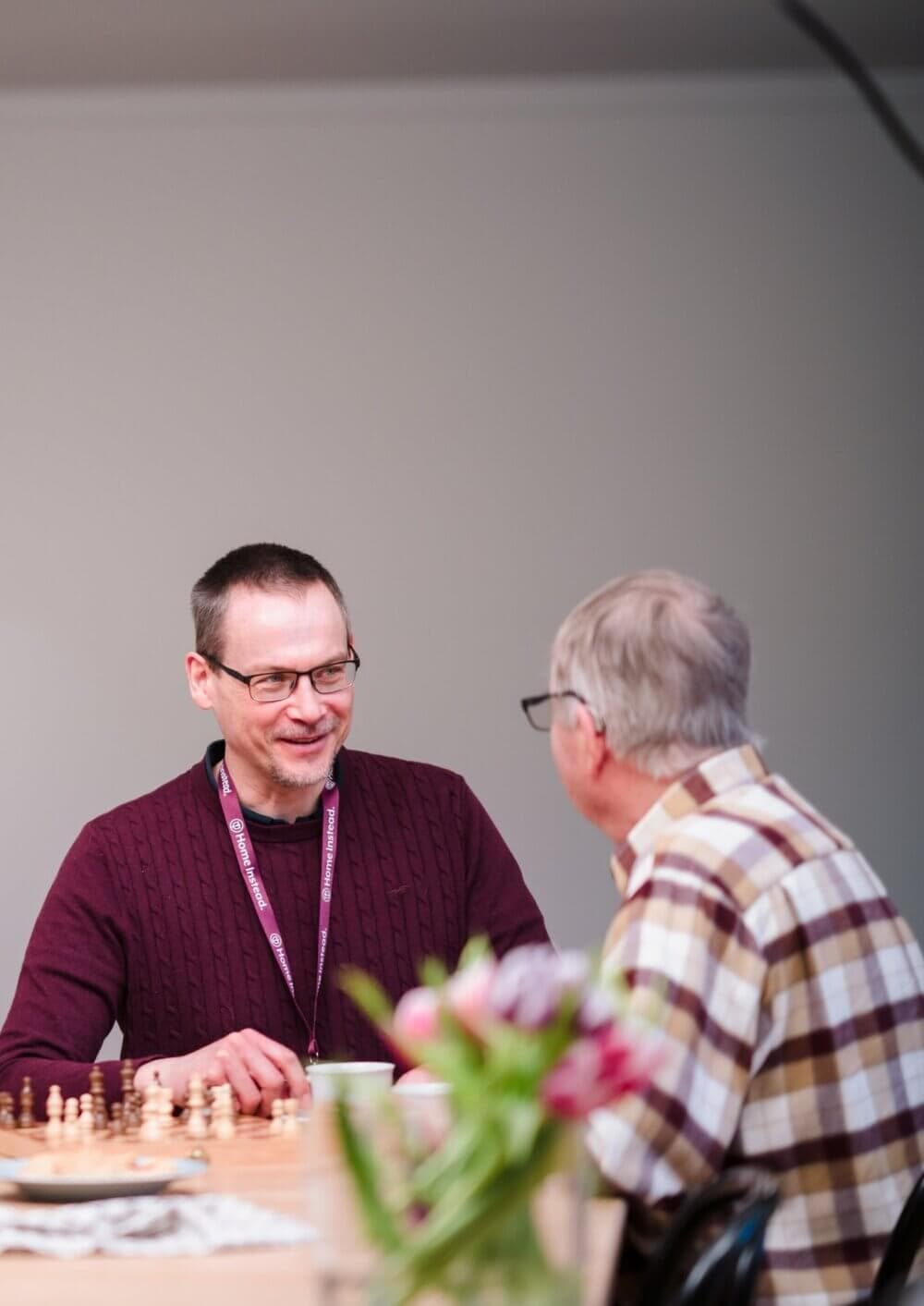 Two men, one wearing a lanyard, sit at a table with a chessboard, engaging in conversation. Flowers are in the foreground. - Home Instead