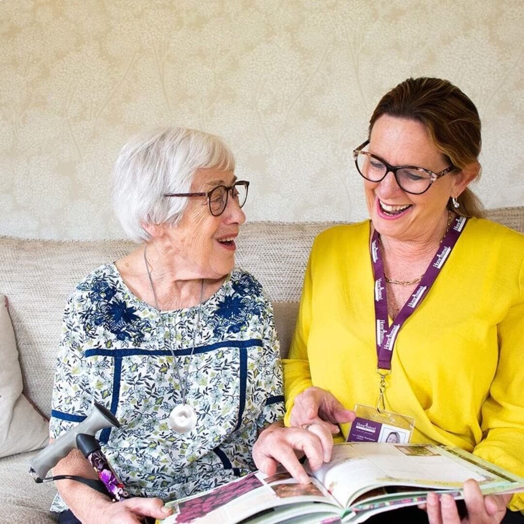 An elderly woman and a caregiver in yellow smile and read a book together on a couch. - Home Instead