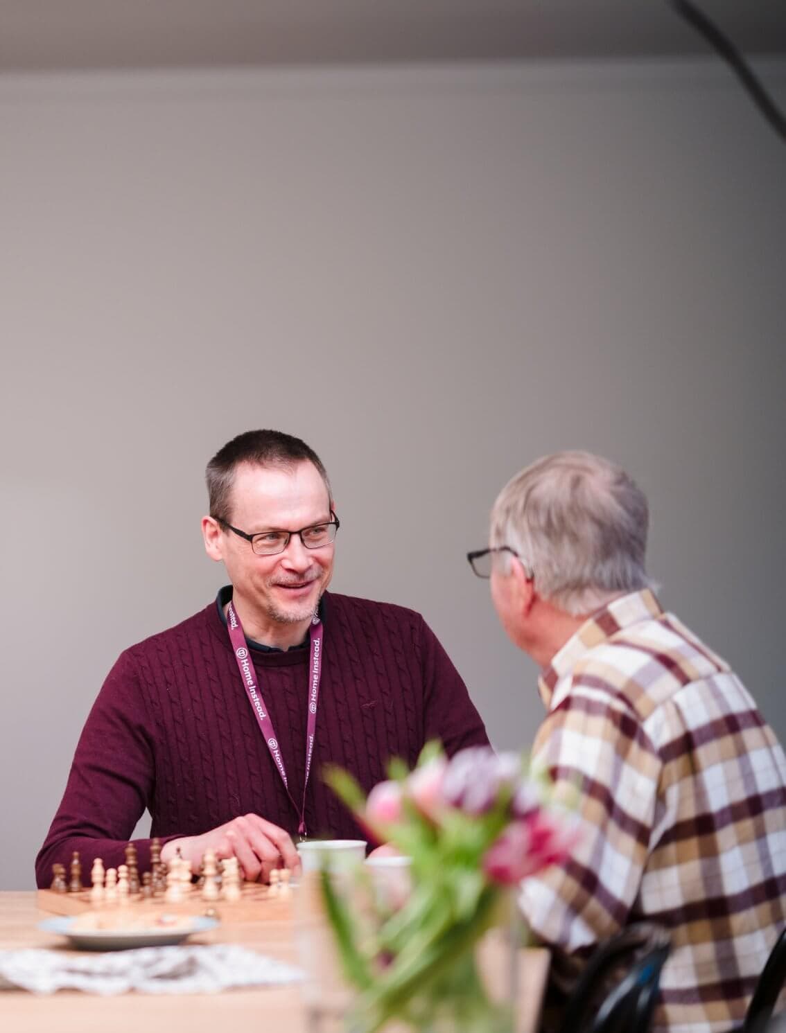 Two men sit at a table, engaged in a conversation with a chessboard in front of them and flowers in the foreground. - Home Instead