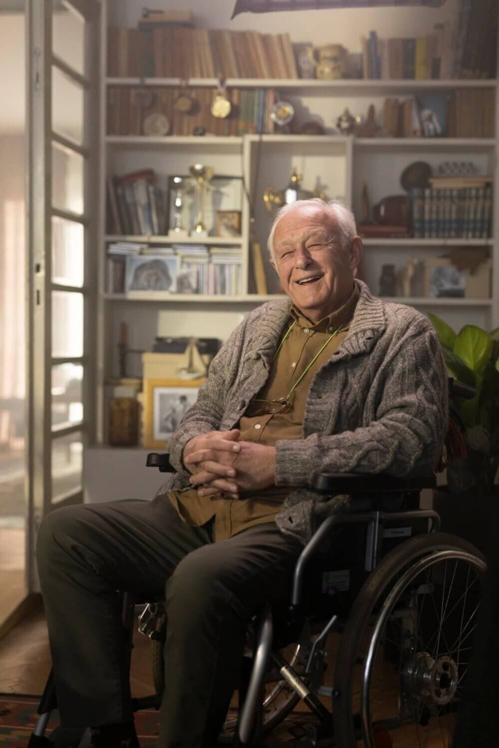 Elderly client in a wheelchair smiling, seated in a cosy room with books and memorabilia on shelves behind him. - Home Instead Bournemouth & Christchurch
