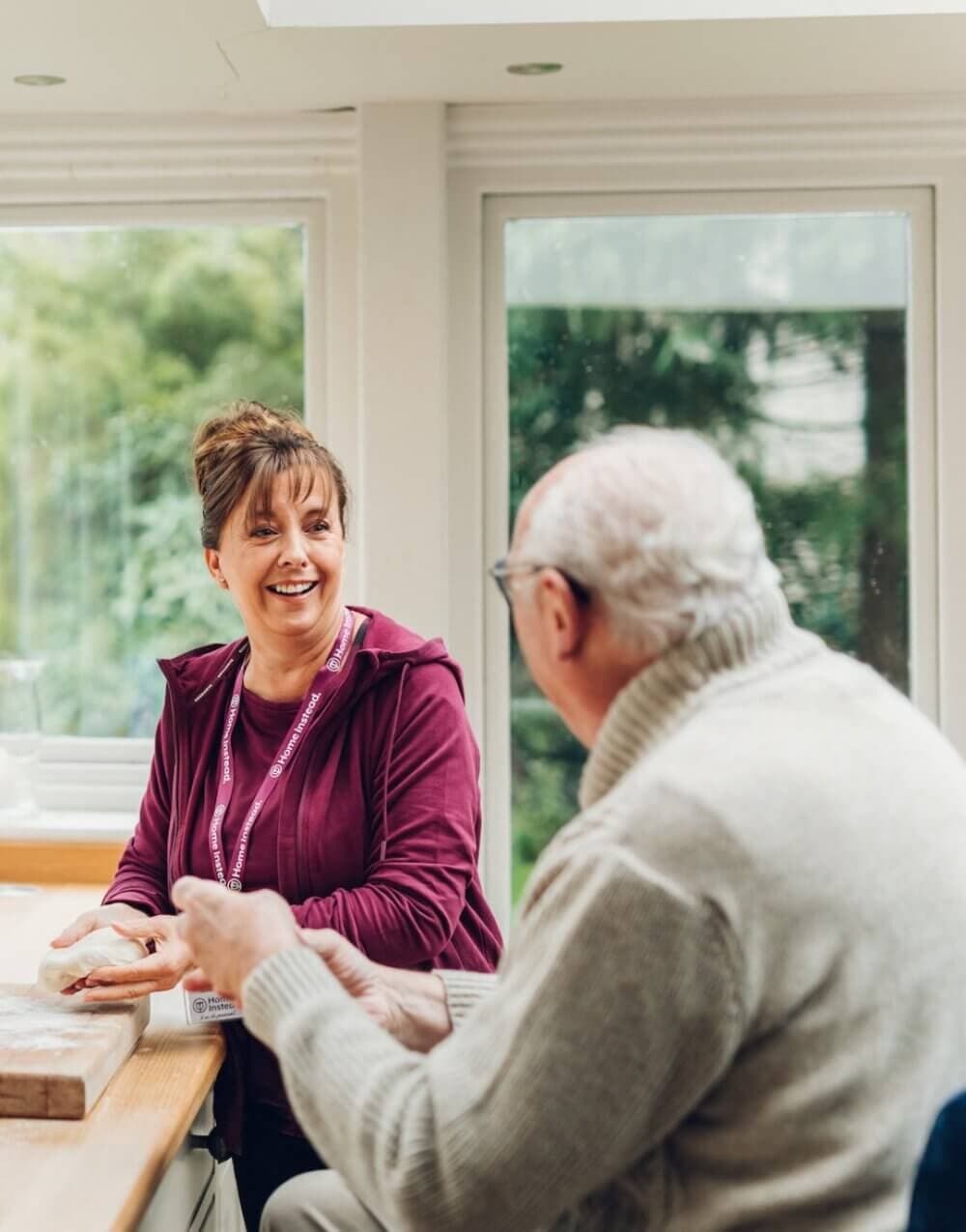 A caregiver in a maroon sweater talks and smiles with an elderly man in a white sweater at a table near large windows. - Home Instead