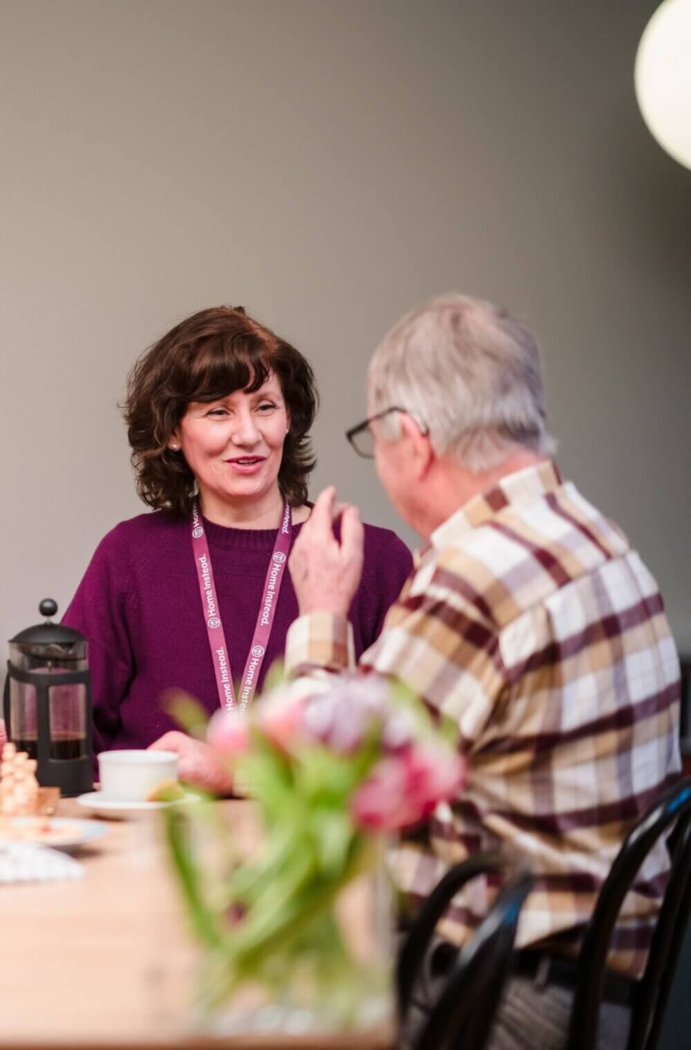 Two people conversing at a table, one wearing a lanyard and the other in a plaid shirt, with a coffee press nearby. - Home Instead