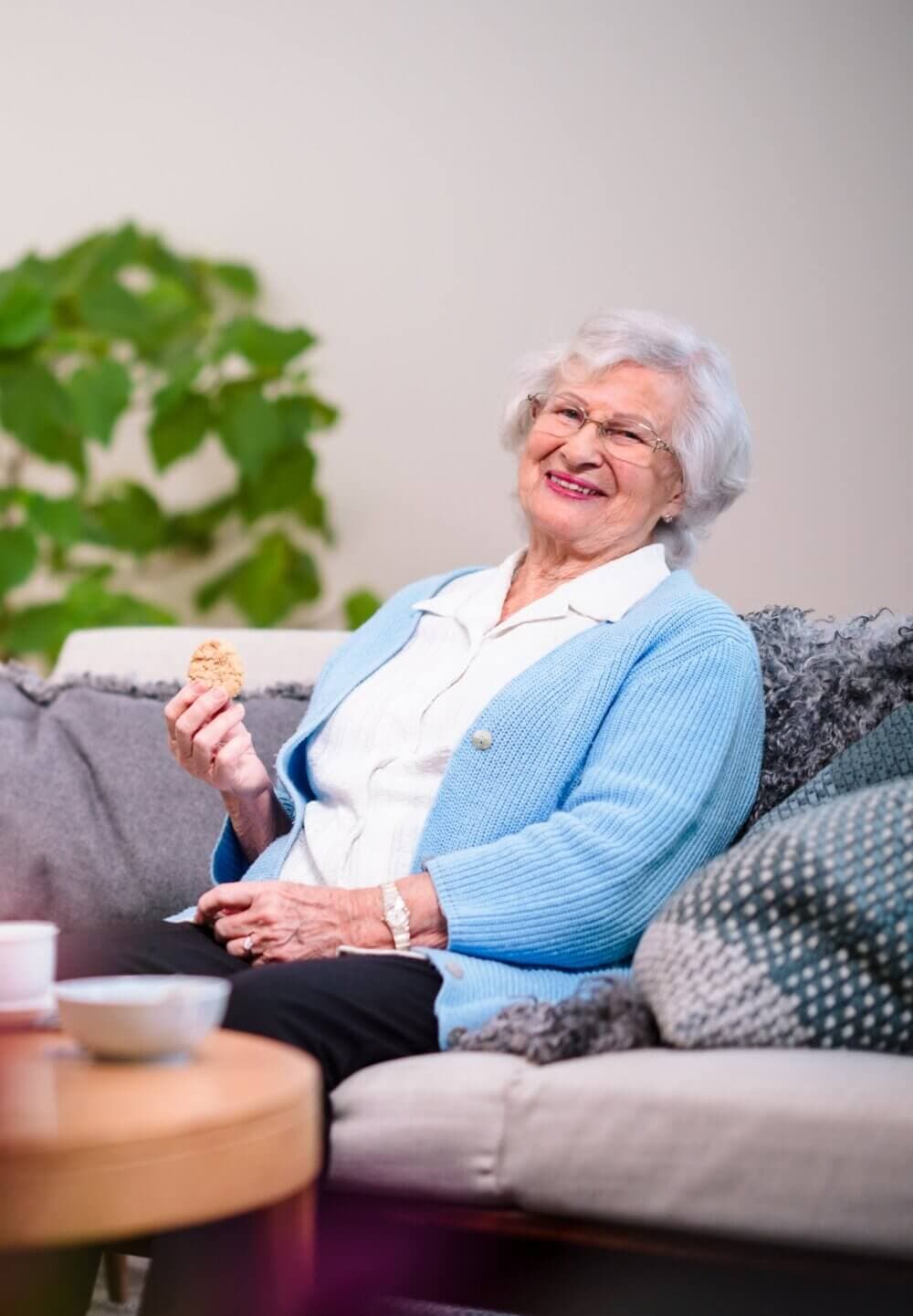Elderly woman with white hair and glasses, smiling while holding a cookie, seated on a couch with greenery in the background. - Home Instead Poole