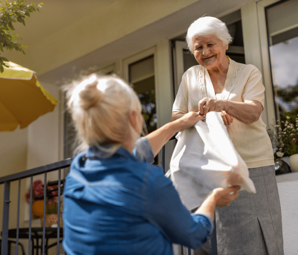 A smiling elderly woman receives a bag from a younger woman outside a house with plants on the windowsill. - Home Instead