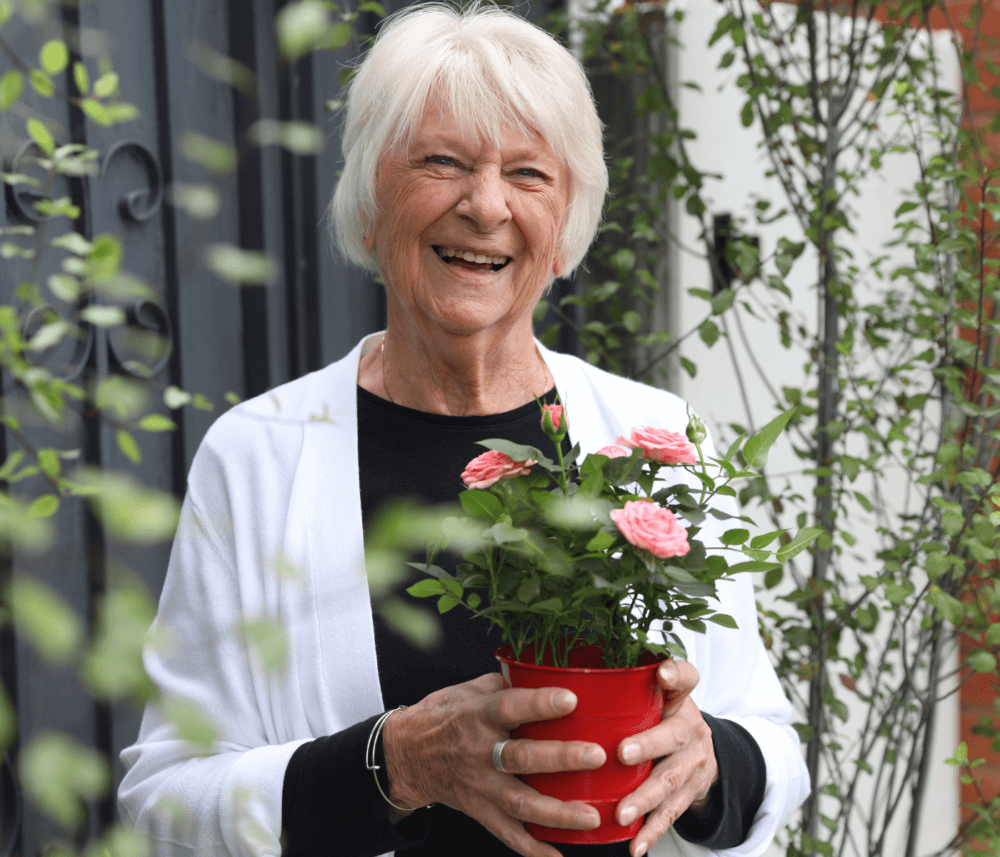 Elderly woman smiling and holding a red pot with pink flowers, surrounded by greenery. - Home Instead