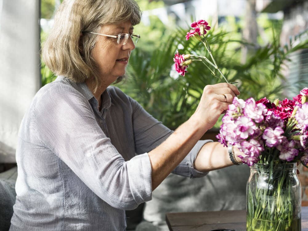 An elderly woman arranges fresh pink flowers in a glass vase, surrounded by greenery. - Home Instead