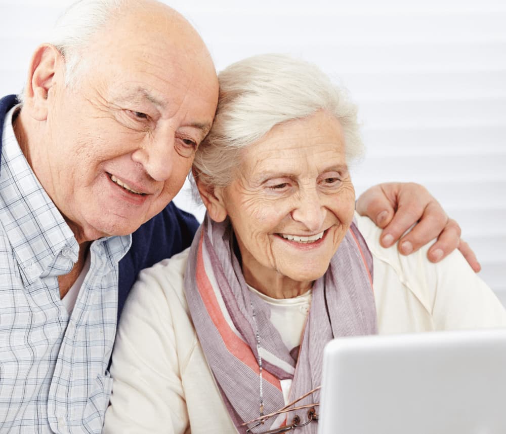An elderly couple smiles as they look at a tablet together, sitting closely and enjoying their time. - Home Instead