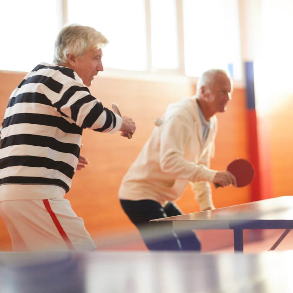 Two elderly men playing a lively game of table tennis indoors, both focused on the ball. - Home Instead