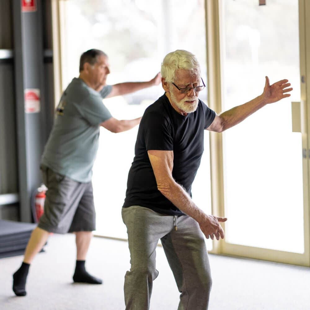 Two elderly men practicing Tai Chi indoors, with one in the foreground and the other in the background near a window. - Home Instead