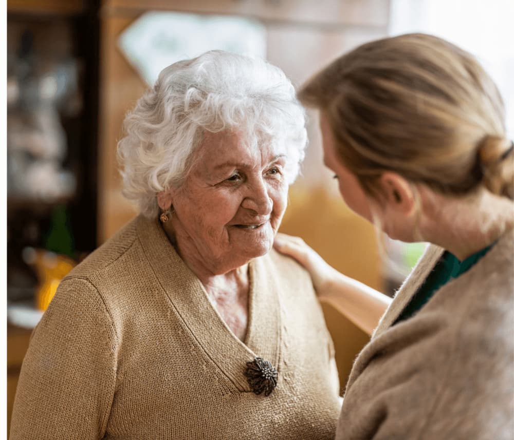 An elderly woman with white hair smiles while talking to a younger woman who is touching her shoulder gently. - Home Instead