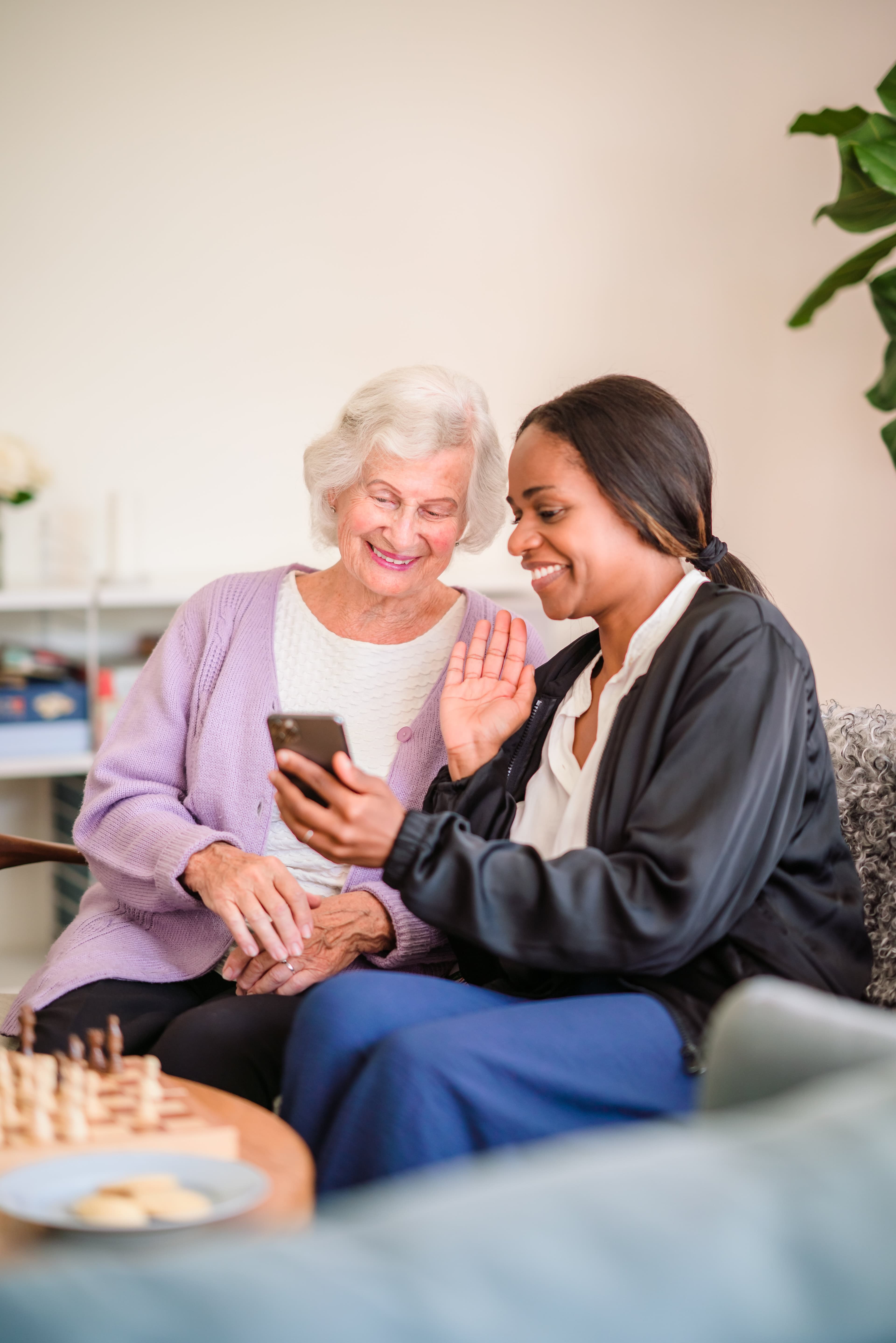 Elderly woman and younger woman smiling, looking at a smartphone, with a chessboard on the table in front of them. - Home Instead