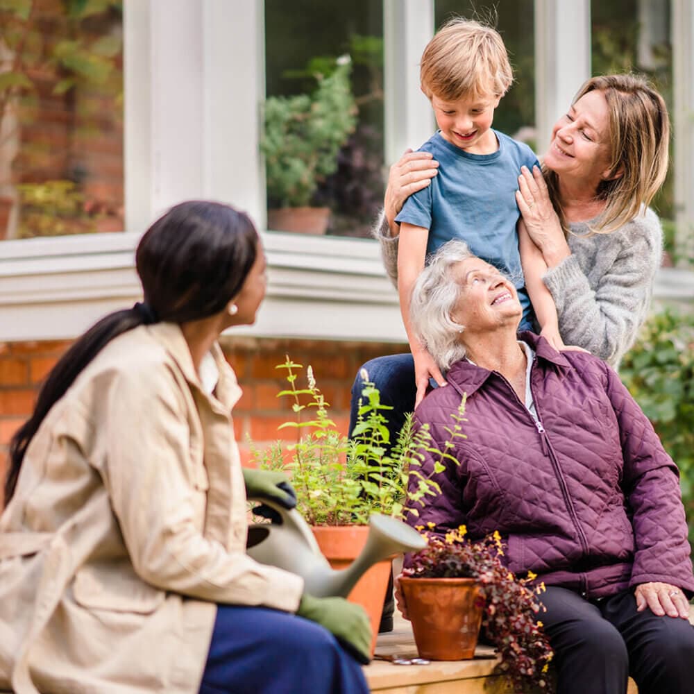 Four people, including a young boy and elderly woman, enjoying a conversation outside a house near plants and flowers. - Home Instead