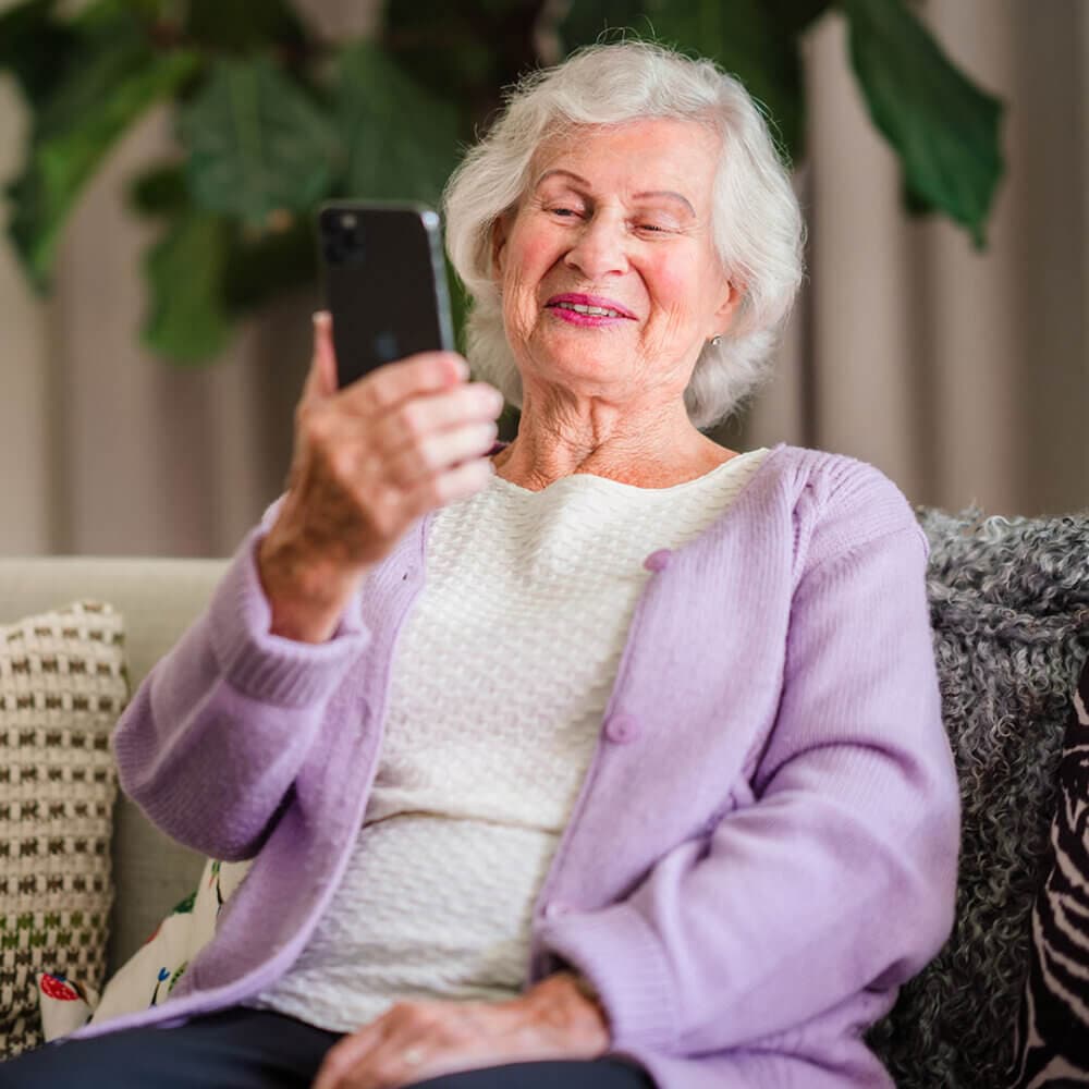An elderly woman with white hair smiles while holding a smartphone and sitting on a couch. - Home Instead