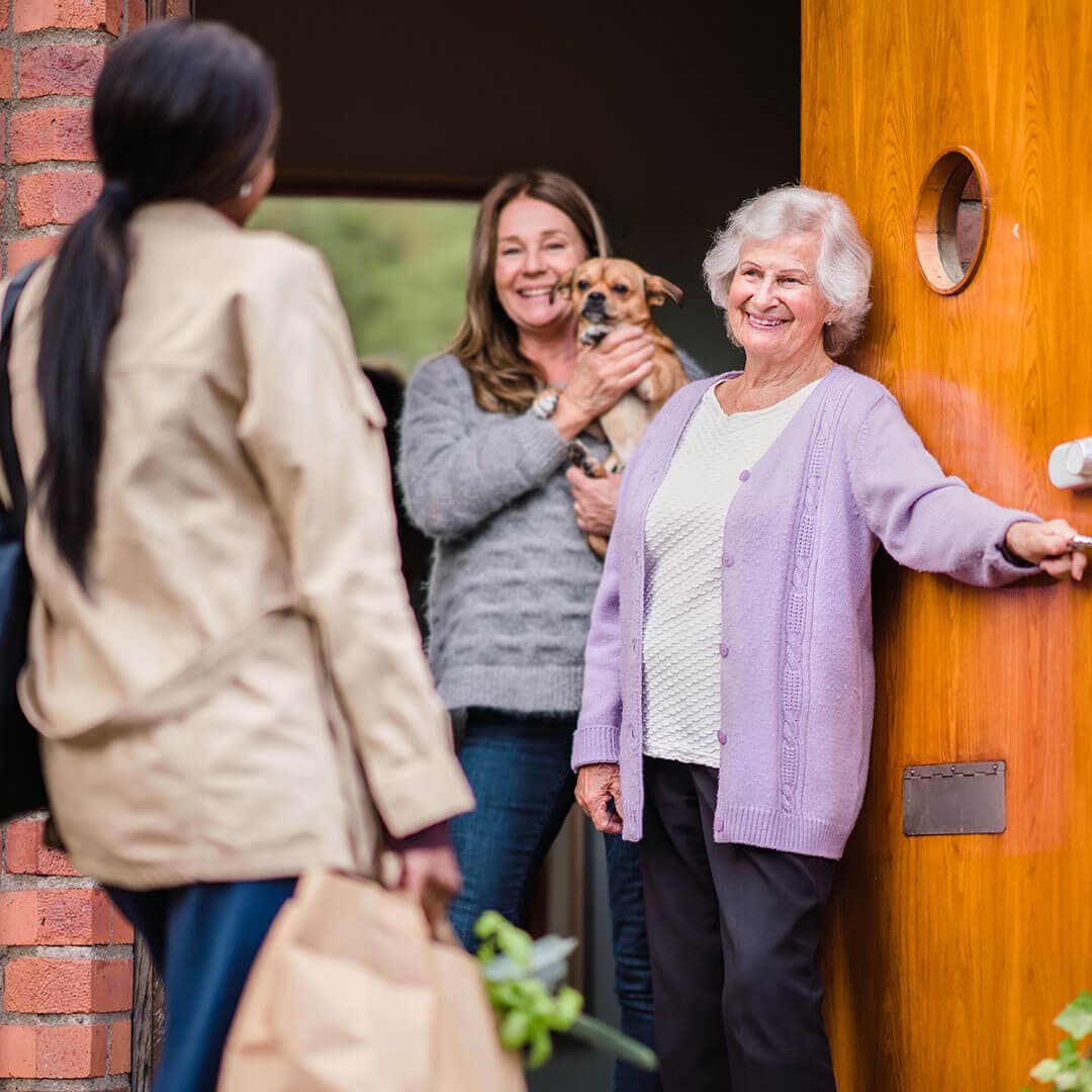 Elderly woman welcomes a guest holding groceries at the door, while another woman inside holds a small dog. - Home Instead