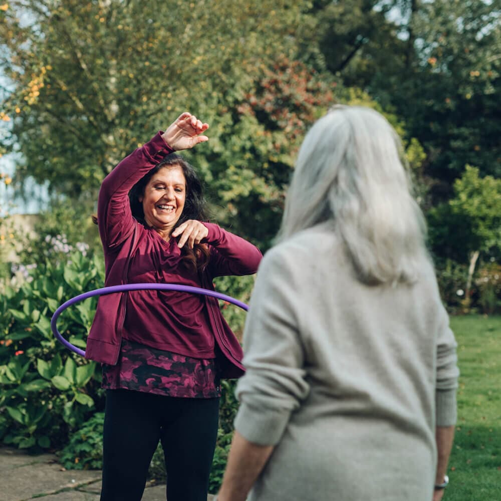 Two women enjoying time outdoors. One woman hula hoops while the other watches and smiles in a garden setting. - Home Instead