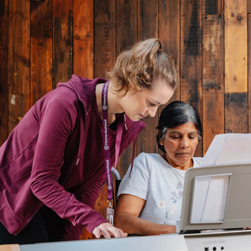 A young woman assists an older woman at a desk with papers. The young woman wears a purple lanyard and cardigan. - Home Instead