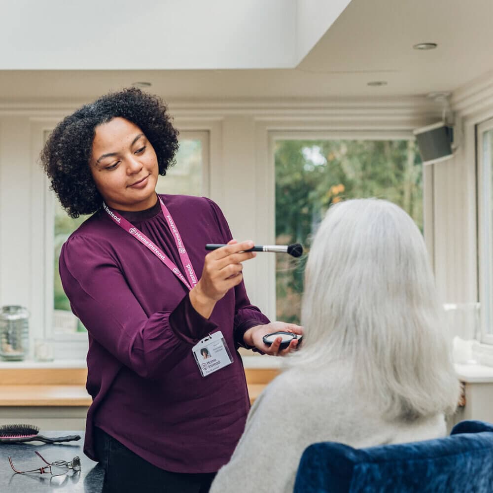 A woman with curly hair applies makeup to an older woman with long white hair in a bright room with large windows. - Home Instead