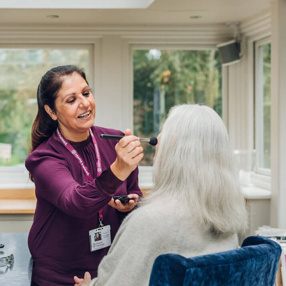 A caregiver in a purple top applies makeup to a seated elderly woman with long white hair in a bright room. - Home Instead