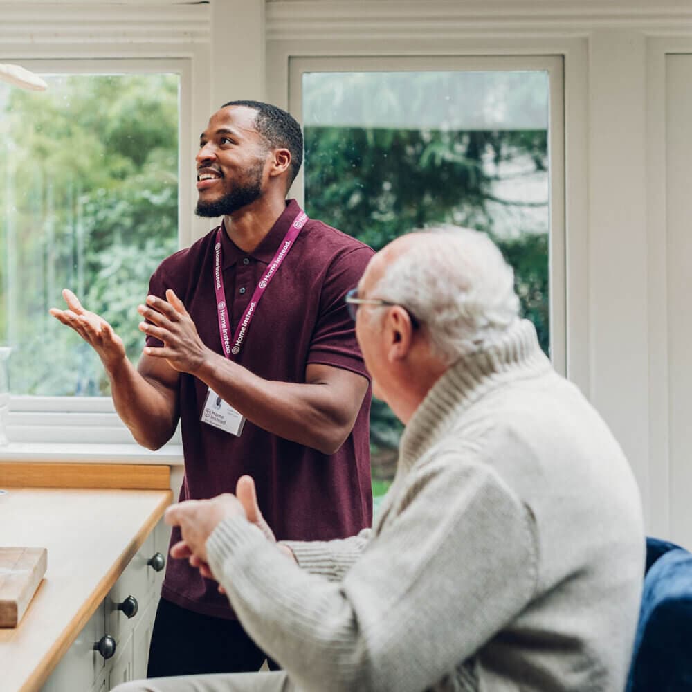 A caregiver in a maroon shirt talks with an elderly man in a sweater, both smiling, in a kitchen with large windows. - Home Instead
