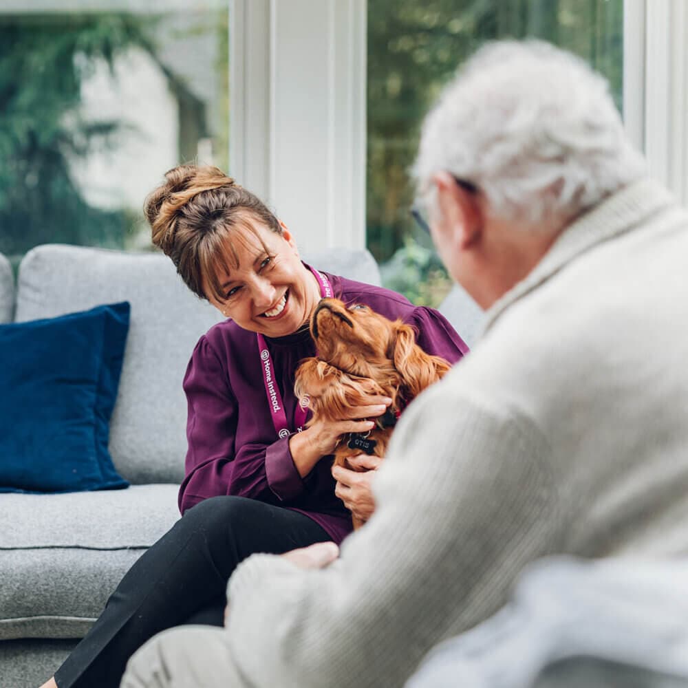 A woman with a dog and an older man wearing a sweater sit together on a couch, smiling and enjoying the moment. - Home Instead
