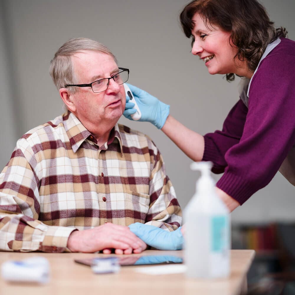 A caregiver in gloves checks an older man's ear using an otoscope, both smiling, hand sanitizer on the table. - Home Instead