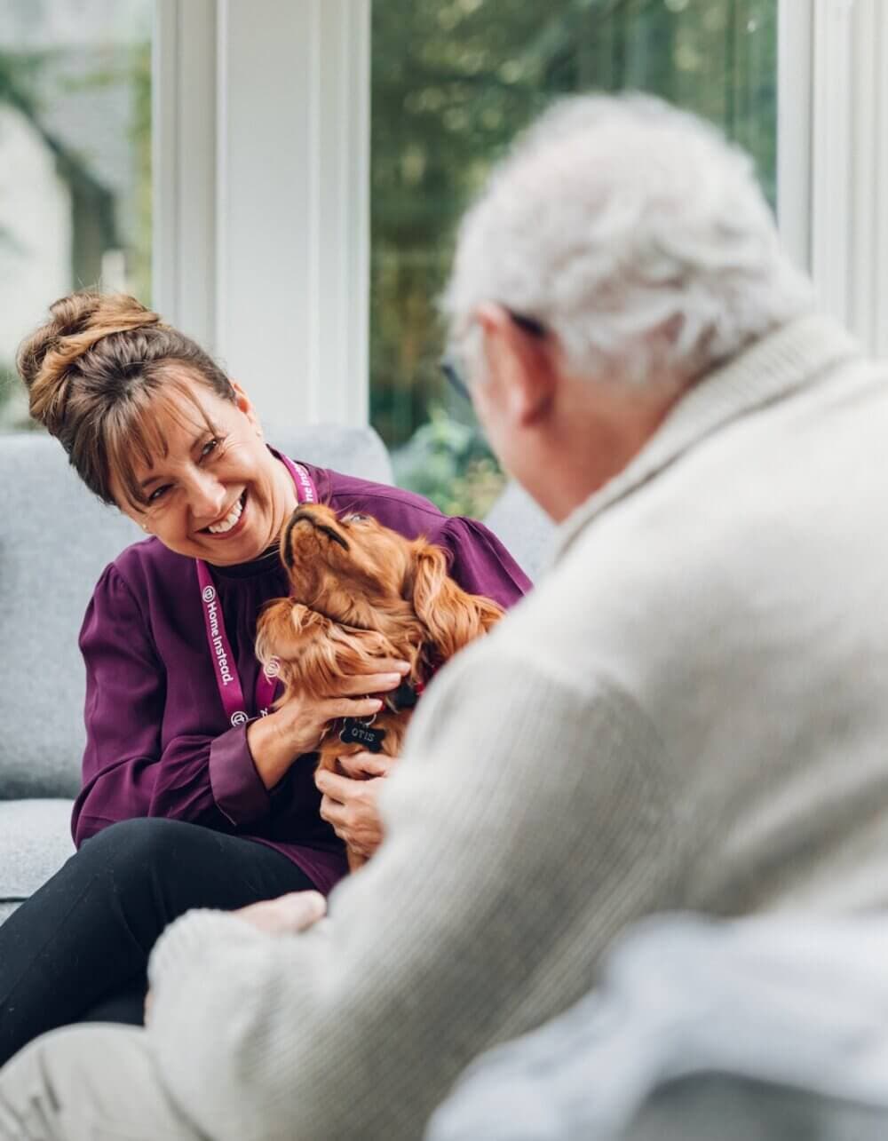 A woman and an elderly man sit on a couch, smiling, as the woman holds a dog looking up at her. - Home Instead