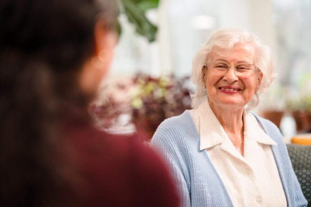 An elderly woman with white hair, wearing glasses and a blue cardigan, smiles while talking to a person with dark hair. - Home Instead