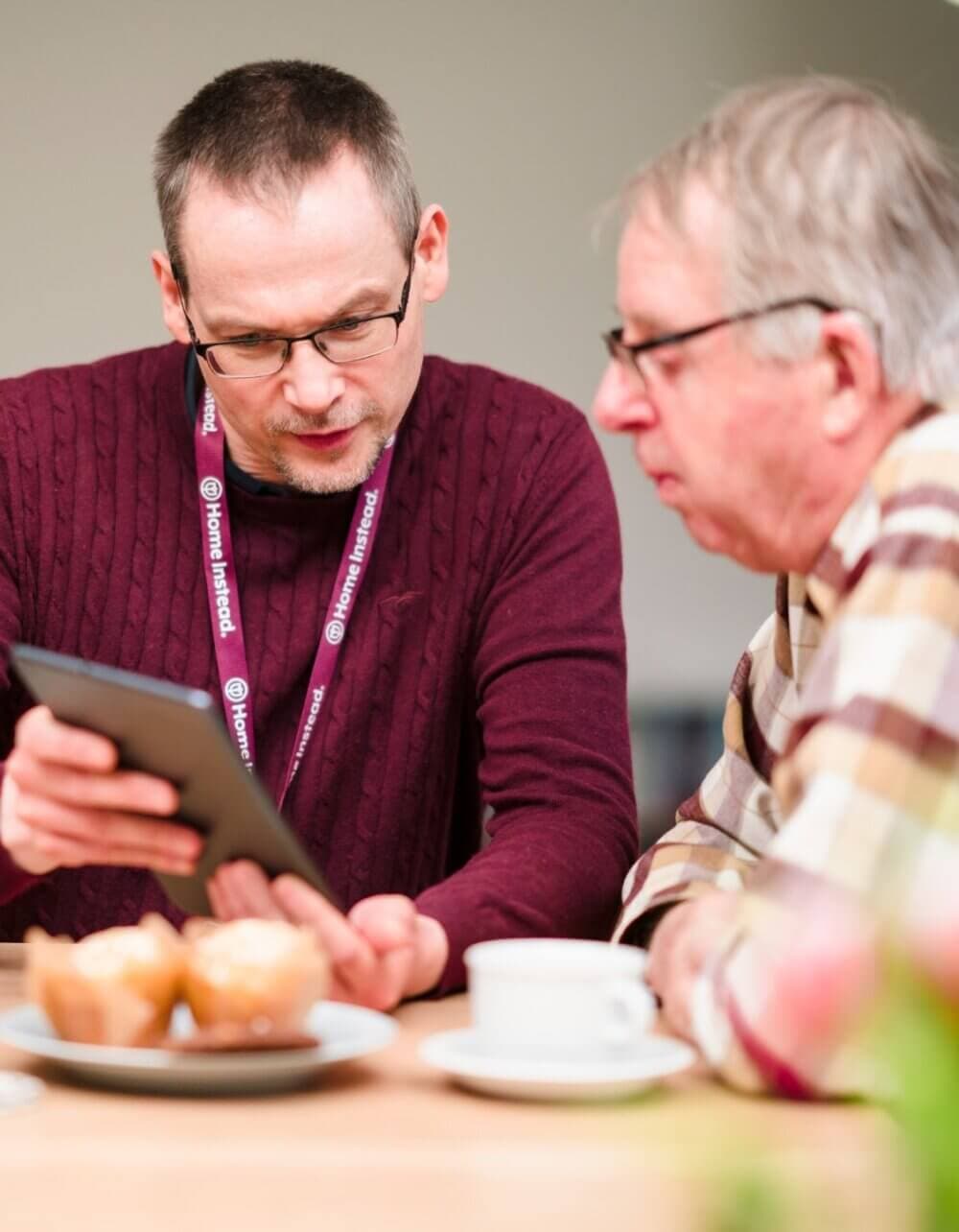 Two men at a table, one is showing the other something on a tablet. There are muffins and a cup on the table. - Home Instead