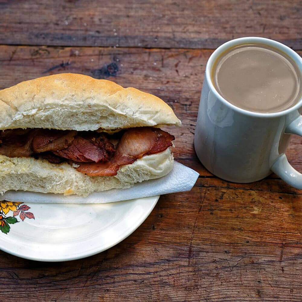 A bacon sandwich on a plate with a mug of coffee on a wooden table. - Home Instead