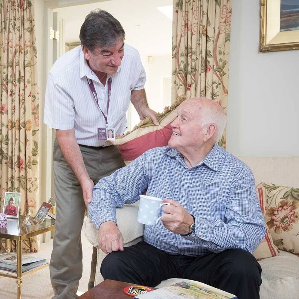 Older man sitting on a couch holding a mug, smiling at another man who is standing and leaning on the couch. - Home Instead