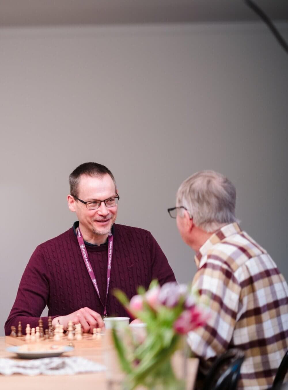 Two people seated at a table with a chess set, smiling and chatting, with a vase of flowers in the foreground. - Home Instead