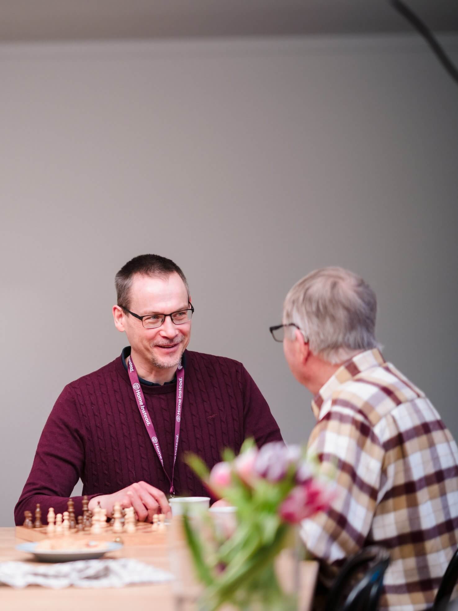 Two men engaged in conversation at a table with a chessboard and flowers in the foreground. - Home Instead