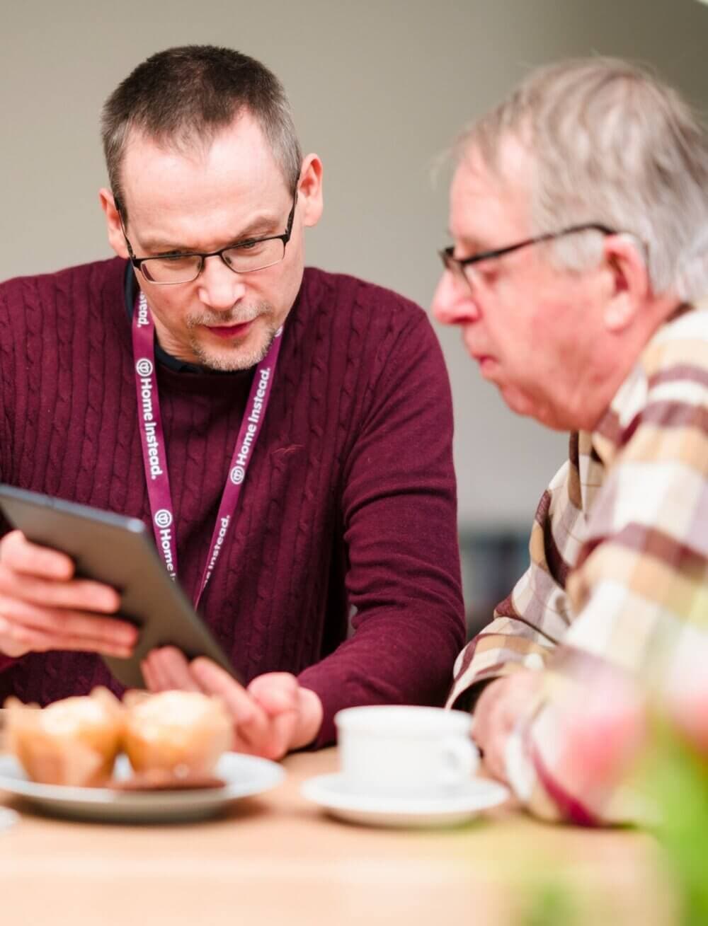 Two men sitting at a table, one holding a tablet, with coffee and muffins in front of them. - Home Instead
