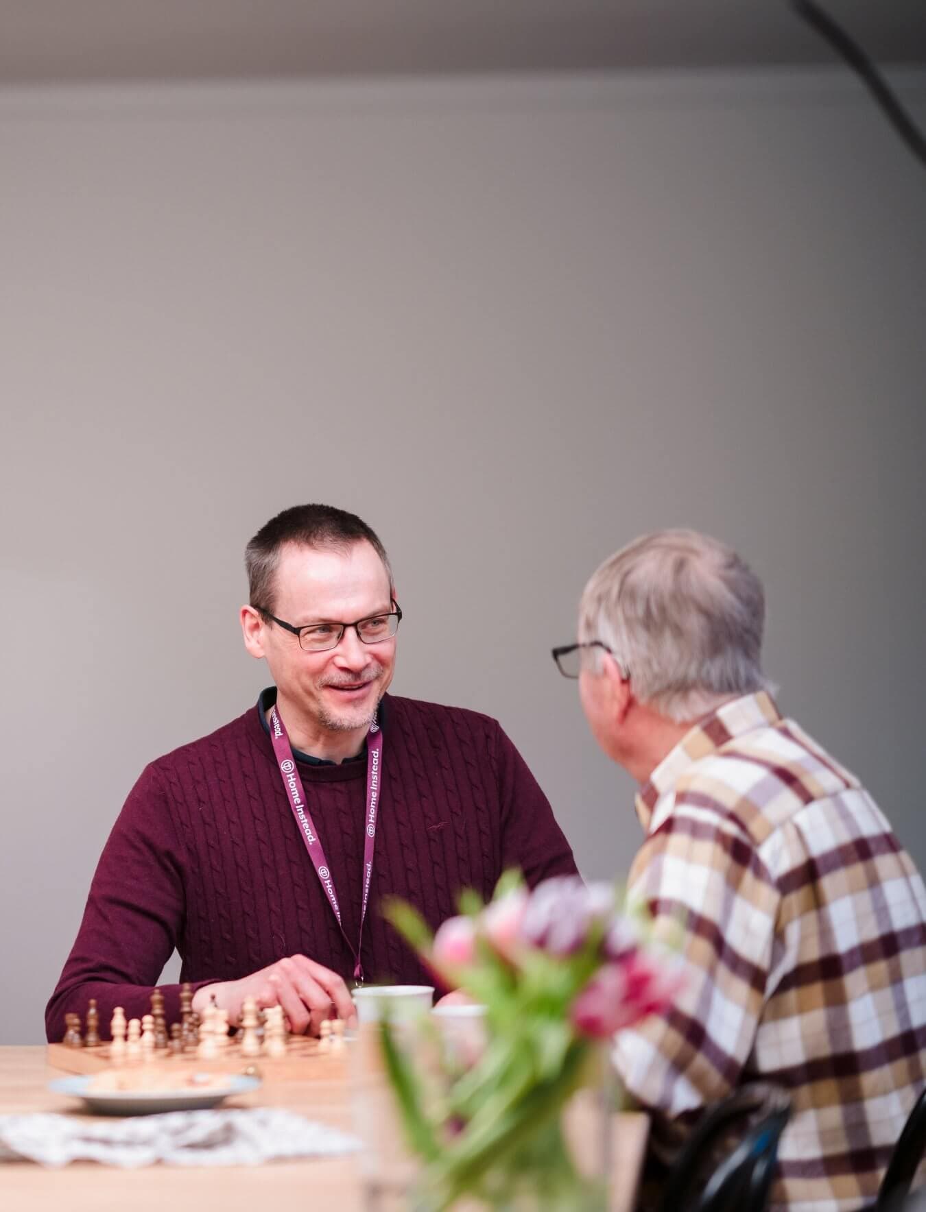 Two men sit at a table chatting, with a chessboard and flowers in the foreground. - Home Instead