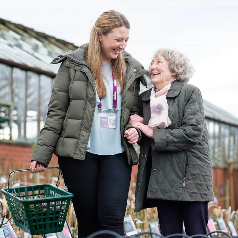 A young woman and an elderly woman smile and walk together, the young woman holds a green basket. - Home Instead